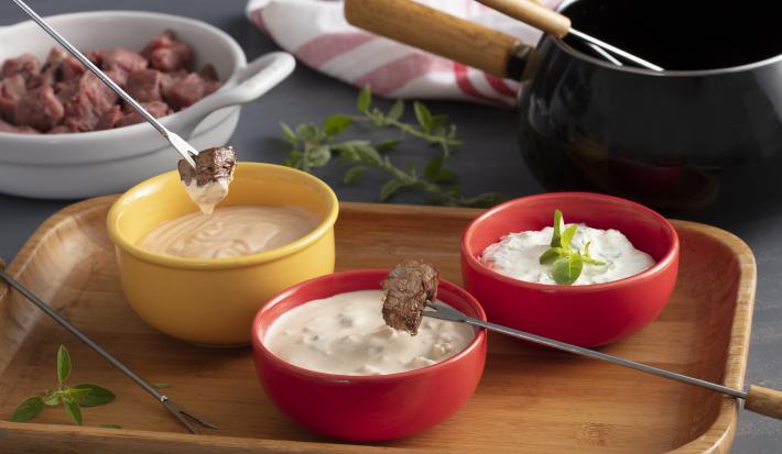 Fotografia em tons de vermelho e amarelo de uma bancada. A frente 1 pote amarelo com molho e 2 potes vermelhos com molho, 2 espetos com pedaços de carne na ponta e ao fundo uma travessa com pedaços de carne crua e uma panela de fondue preta.