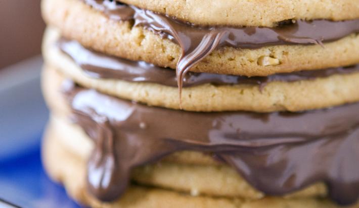 Fotografia de biscoitos empilhados, e entre alguns, um creme de chocolate caindo um pouco nas bordas. Os biscoitos estão em um prato quadrado na cor azul em tom mais escuro, que estão sobre uma mesa na cor marrom escuro.