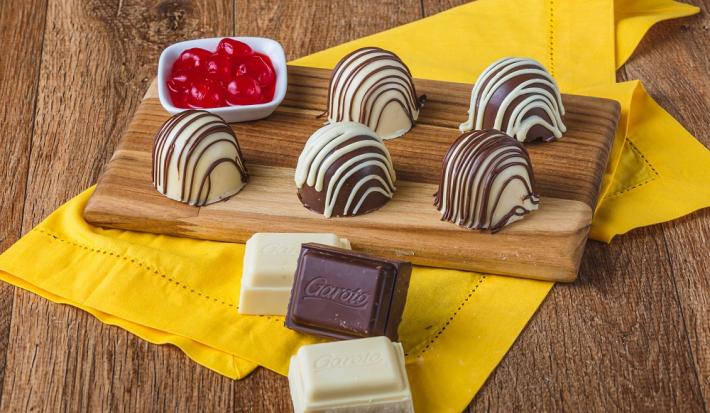 Fotografia em tons de marrom em uma bancada de madeira de cor marrom. Ao centro, um pano amarelo contendo uma tábua de madeira com as trufas e ao lado, há um recipiente contendo cerejas.