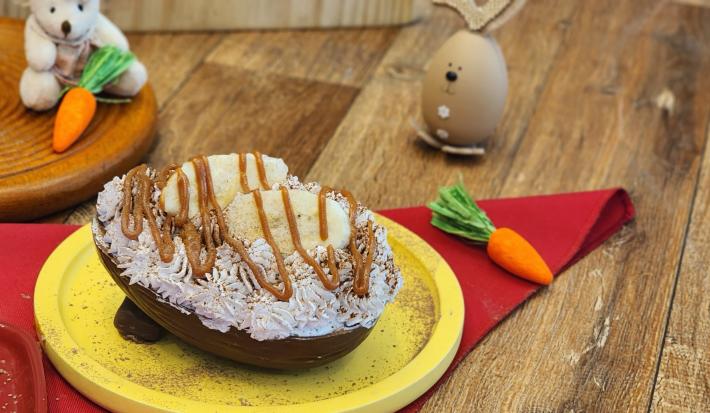 Fotografia em tons de amarelo e vermelho em uma mesa de madeira com um prato amarelo ao centro. Dentro dele existe um ovo de chocolate recheado e coberto com chantilly, doce de leite e banana. Ao fundo tem um coelho e algumas cenouras de pelúcia.