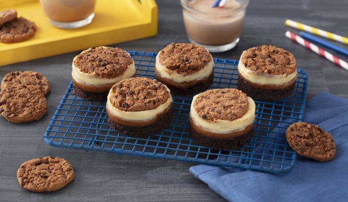 Foto de uma bancada cinza com uma grade contendo cinco doces circulares em camadas diferentes e decorados com cookies Nescau. Além disso há um tecido azul à direita e, ao fundo, canudos coloridos de papel, uma bandeja amarela e copos com leite e Nescau