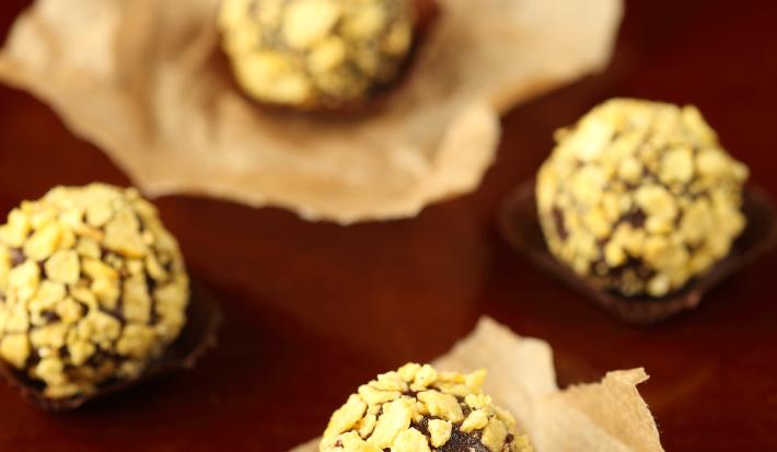 Fotografia de cinco brigadeiros enrolados e passados em nozes, alguns em forminhas, outros sobre um papel manteiga. Ao fundo, forminhas empilhadas, sobre uma mesa marrom em tom escuro.