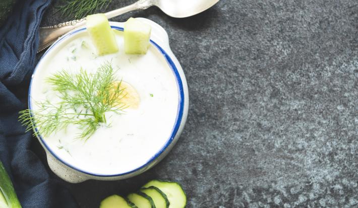 Fotografia vista de cima de um recipiente em vidro na cor branca com bordas na cor azul. Dentro, um caldo de pepino, com decorações do fruto dentro e ao lado, também. Ao lado, uma colher de sopa, sobre uma mesa de mármore em tom escuro.
