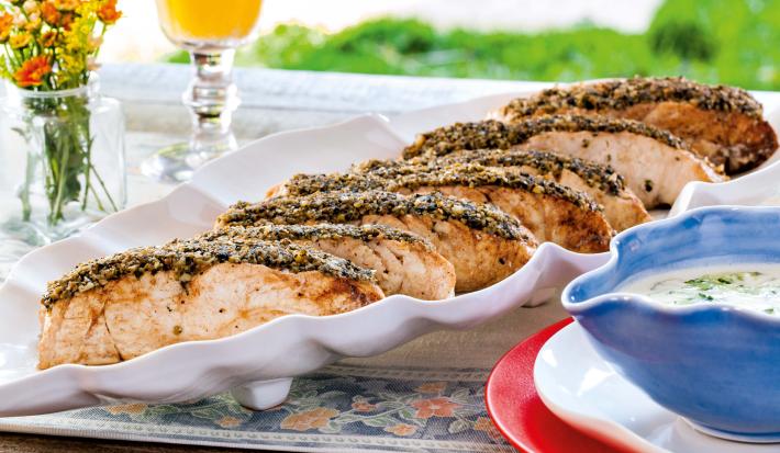 Fotografia em tons de laranja em uma mesa de madeira com um recipiente branco ao centro com as postas de robalo com crosta de grãos em cima dele. Ao lado, um prato vermelho e um potinho azul em cima com o molho de iogurte. Ao fundo, uma taça com suco.