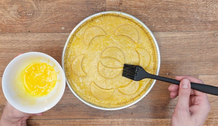 Mão pincelando gema de ovo sobre superfície de torta decorada com meias luas de massa.