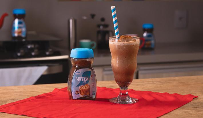 Fotografia de um copo de vidro grande com frozen de café, ao lado de uma embalagem de nescafé gelado, sobre um pano vermelho e uma mesa de madeira. Dentro do copo tem um canudo azul e branco apoiado.