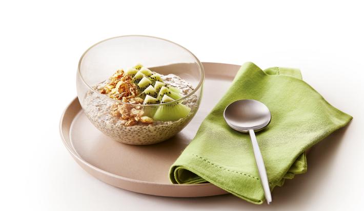 Foto da receita de Overnight Oats de Kiwi. Observa-se um prato de cerâmica marrom com um recipiente de vidro em cima com a receita. Ao lado direito, um guardanapo verde com uma colherinha.