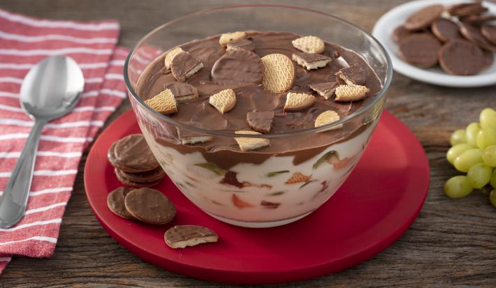 Foto da receita de Taça Cremosa de Calipso, feita em uma taça de vidro grande, sobre um prato vermelho com alguns biscoitos ao redor, sobre uma bancada de madeira decora com um guardanapo de tecido e com algumas uvas verdes ao lado