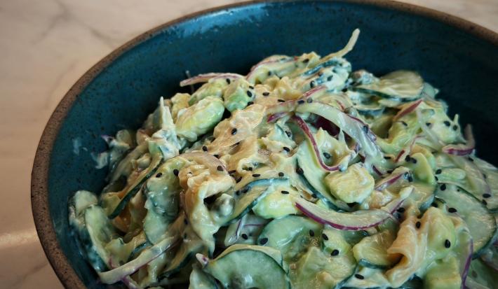 Fotografia em tons de marrom escuro com um bowl escuro ao centro. Dentro do bowl existe uma receita de salada de pepino feita com cream cheese, salmão, cebola e gergelim preto