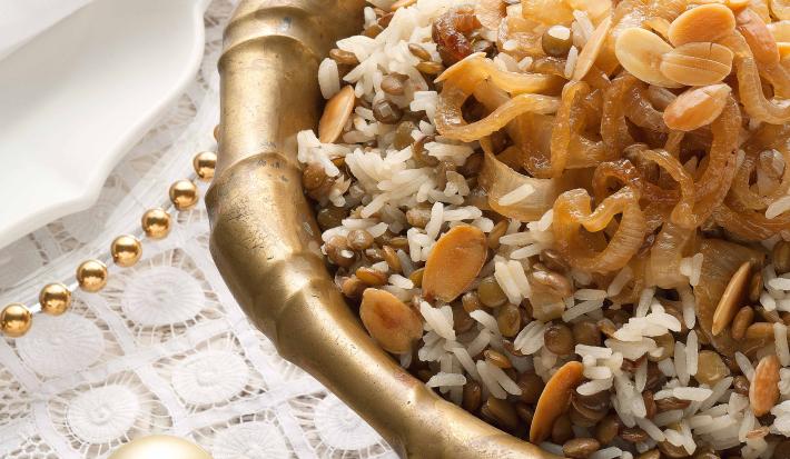 Fotografia em tons de marrom e dourado em uma mesa de madeira com uma toalha branca de renda com um recipiente redondo fundo e grande dourado com o arroz de festa com lentilhas.