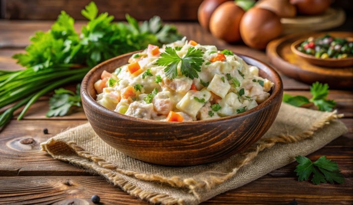 Em cima de uma mesa, travessa com salada de maionese com cenoura, batata, presunto e ervilha. Decorado com folhas de salsa.