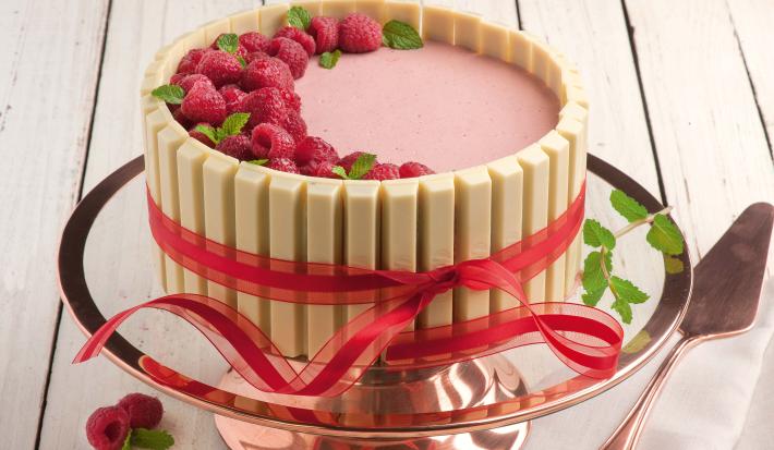 Fotografia em tons de rosa em uma bancada de madeira clara, um suporte para bolo em vidro rosado, com a mousse de framboesa com KitKat Branco ao redor, formando uma linda torta.