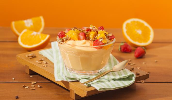 Fotografia em tons de laranja em uma bancada de madeira, uma tábua de madeira, um paninho verde, um recipiente de vidro com o iogurte de laranja, morango e granola. Ao fundo, laranja e morango.