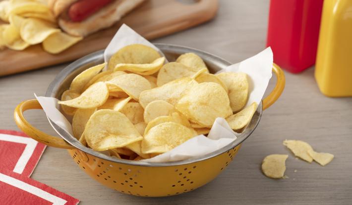 Fotografia em tons de vermelho em uma bancada de madeira clara, um pano vermelho com listra branca, uma tábua de madeira com um cachorro-quente e batatas chips ao lado. Ao centro, um recipiente redondo amarelo com muitas batatas chips.