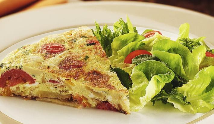 Fotografia em tons de verde em uma bancada com um prato branco redondo raso, um pano amarelo de fundo e a tortilla de omelete com batata e tomate em cima dele, ao lado, salada de alface e tomate cereja.