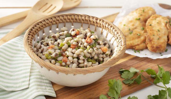 Fotografia em tons de verde em uma bancada de madeira clara, um pano verde listrado, uma tábua de madeira com um recipiente branco com detalhes beges redondo fundo com a salada do feijão-de-corda com vegetais dentro dele.