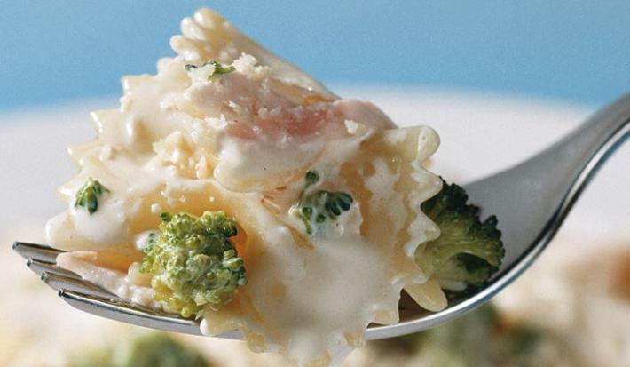 Fotografia em tons de azul, branco e verde, ao centro porção de macarrão com brócolis ao creme suspenso em um garfo, abaixo prato branco com macarrão, creme e brócolis, tudo sobre bancada azul.
