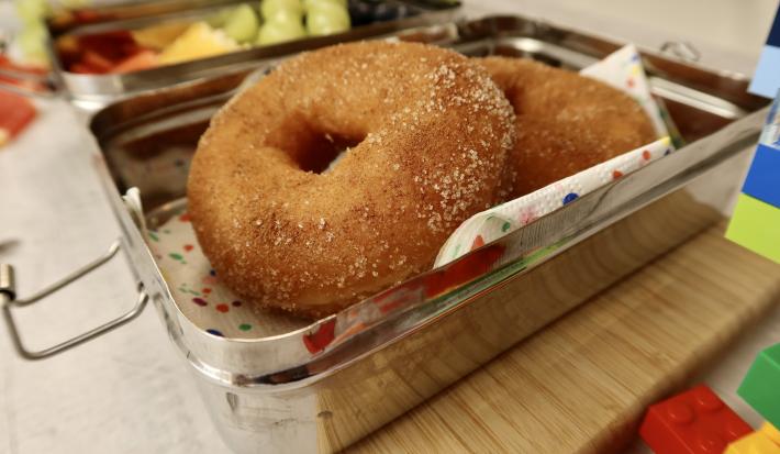 Fotografia em tons de vermelho em uma bancada de madeira clara, uma marmita ao meio com dois donuts de maçã dentro. Ao lado, brinquedos infantis.