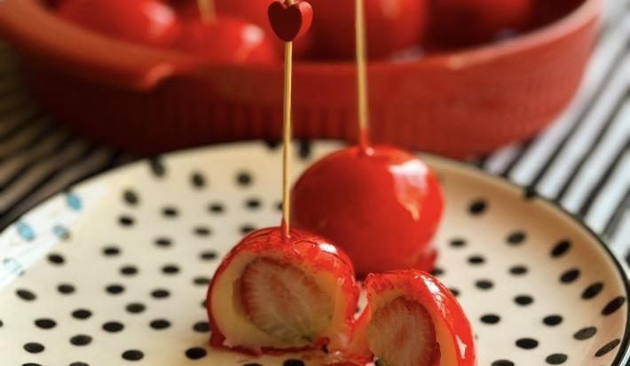 Fotografia de dois morangos do amor prontos em um prato raso branco com bolinhas pretas. Um dos morangos está cortado na metade, mostrando o interior. Ao fundo tem mais porções dentro de um recipiente grande vermelho.
