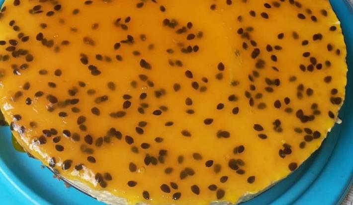 Fotografia em tons de amarelo, em uma bancada de madeira de cor branca. Ao centro, um prato azul contendo a torta de maracujá.