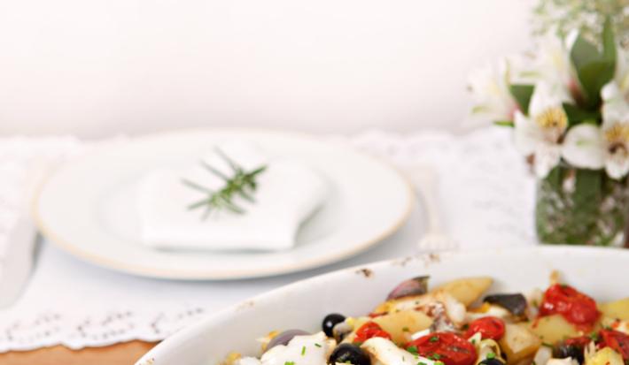 Fotografia em tons de branco e vermelho de uma bancada de madeira vista de lado, ao centro um recipiente oval branco, dentro dele pedaços de bacalhau, tomates cereja e azeitonas. Ao fundo um prato branco e um guardanapo para decorar.