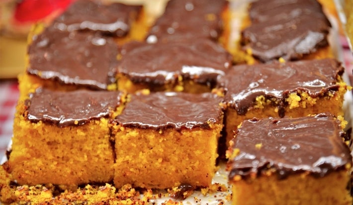 Bolo de cenoura cortado em quadrados, com cobertura de chocolate por cima. A superfície do bolo tem um aspecto mais rústico, com pedacinhos soltos e farelos ao redor. A textura é fofa e bem amarelinha.