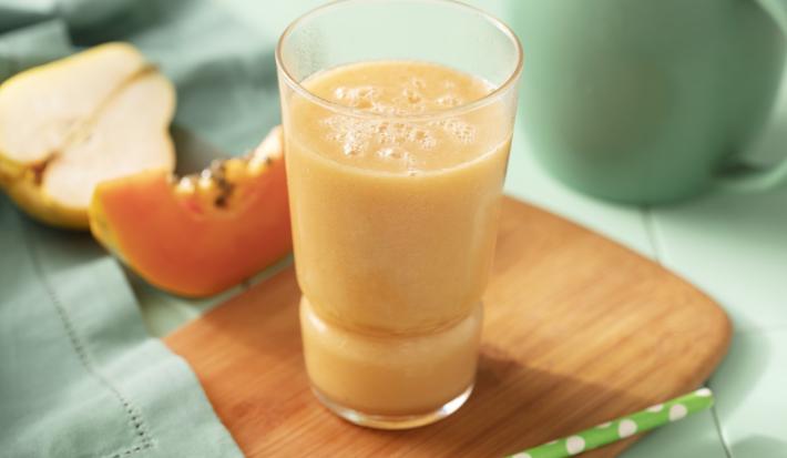 Fotografia em tons de verde e laranja de uma bancada verde, paninho verde, fatia de pera e mamão e uma tábua com um copo com a bebida laranja. Ao fundo uma jarra verde.