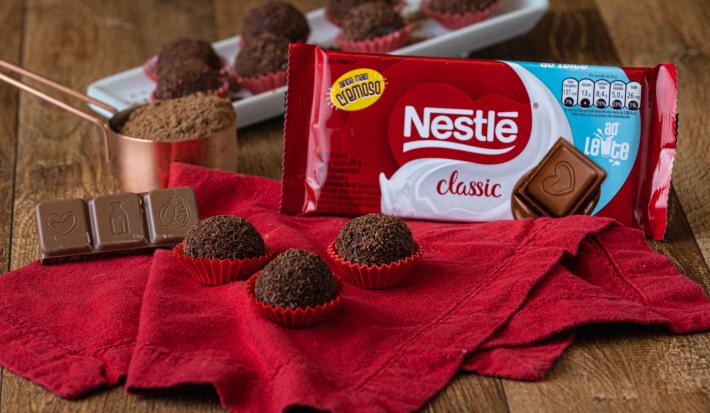 Foto aproximada da receita de brigadeiro gourmet, em uma bancada de madeira decorada com um tecido vermelho, uma barra de chocolate ao leite e mais brigadeiros sobre um prato branco