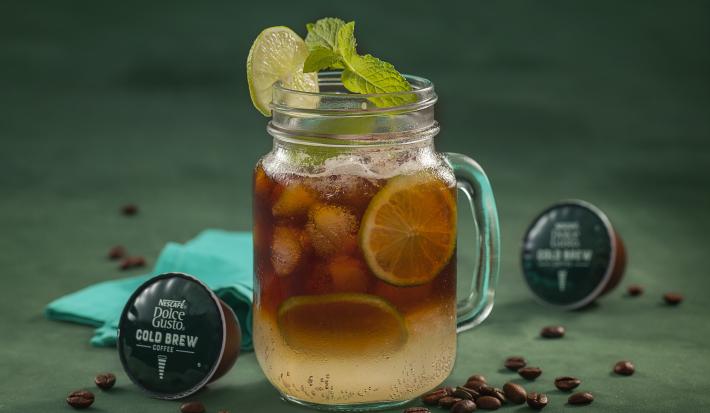 Fotografia em tons de verde de uma bancada verde, sobre ela um copo de vidro com cold brew e limões. Ao lado capsulas de Dolce Gusto Cold Brew e um paninho azul.