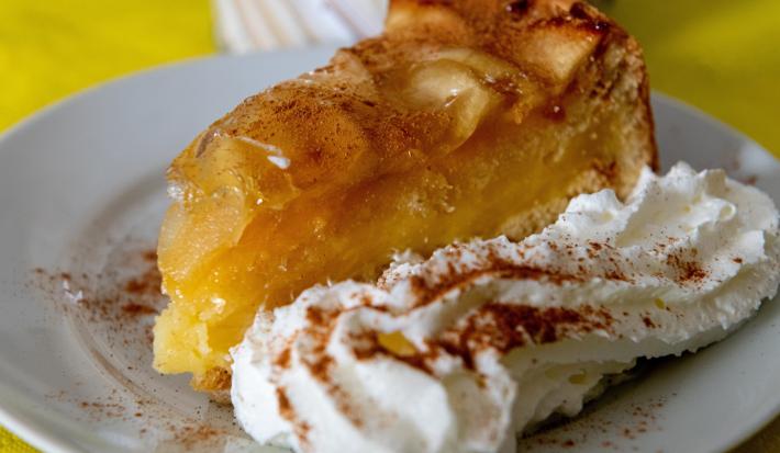 Fotografia em tons de branco e amarelo com um prato branco ao centro. Dentro do prato existe um bolo de maçã polvilhado com canela acompanhado de um jato de chantilly.