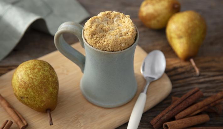 Fotografia em tons de amarelo em uma bancada de madeira de cor marrom. Ao centro, uma tábua de madeira de cor amarela, em cima há uma caneca contendo o bolo e ao lado, há uma pera e uma colher pequena. Ao fundo, há 2 peras e um pano verde claro.