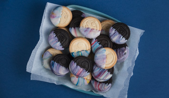 Biscoitos claros e escuros com cobertura marmorizada, dispostos sobre papel manteiga dentro de prato azul, vistos de cima.