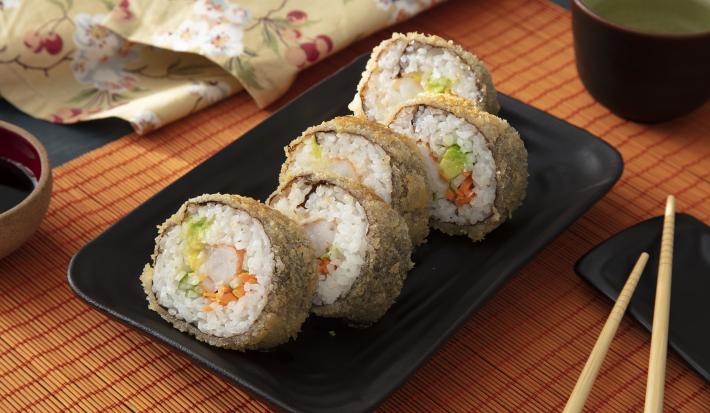 Foto da receita de Hot Roll. Observa-se 5 hot rolls em um prato de cerâmica preto. Ao lado direito, apoiado no prato, hashis. À frente, um potinho com molho shoyu.