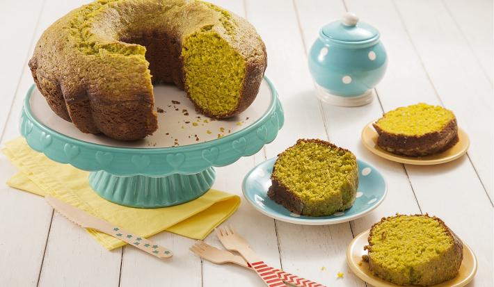 Fotografia em tons de amarelo em uma bancada de madeira de cor branca. Ao centro, um pano amarelo com uma boleira verde em cima contendo o bolo. Ao lado, há 3 pires contendo 3 fatias do bolo e ao fundo, uma chaleira.