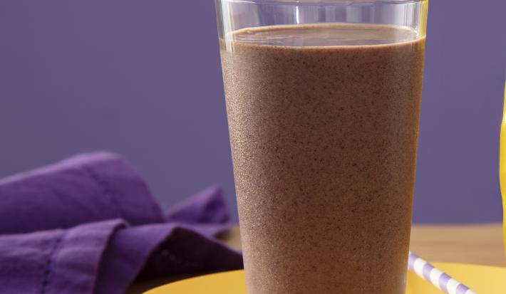 Fotografia de um copo de vidro grande com um smoothie de açaí apoiado em um prato amarelo raso com um canudo branco e roxo. Ao lado tem uma embalagem de ninho proteen, sobre uma mesa de madeira.