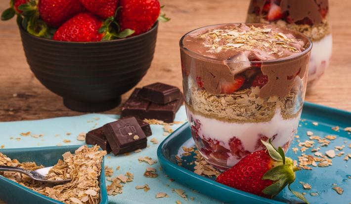 Fotografia em tons de vermelho em uma bancada de madeira, uma tábua azul, um recipiente quadrado azul com um copo de vidro com o creme branco, com Neston 3 Cereais, creme de chocolate e morangos.