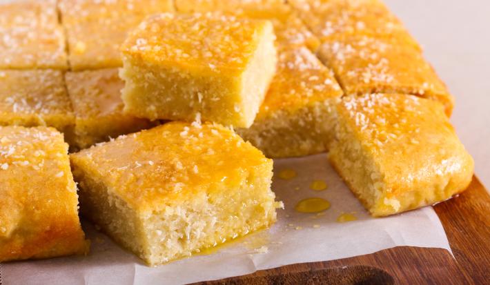 Fotografia em tons de amarelo com uma tábua ao centro. Em cima da tábua existe vários pedaços de bolo de cor amarela ao centro. Os pedaços de bolo estão cobertos com uma calda e coco ralado