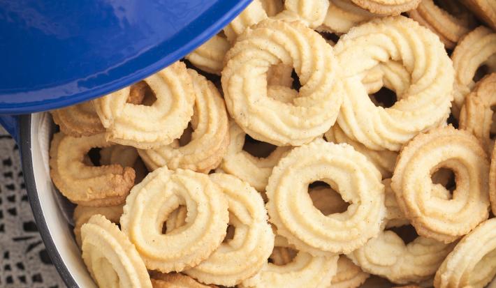 Fotografia em tom claro, com biscoitos amanteigados em formato redondo, modelado com bico de confeitar trançado, dentro de um refratário. No canto superior esquerdo da foto, uma tampa azul.