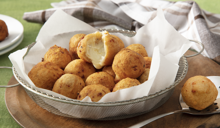 Foto da receita de bolinho de batata servida em diversos bolinho dentro de uma peneira de metal coberta com guardanapos sobre uma tábua de madeira com uma colher prateada ao lado com um bolinho. Ao fundo um paninho cinza quadriculado