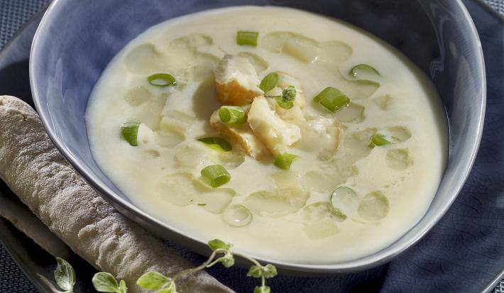 Fotografia de um recipiente azul de vidro com uma sopa de alho poró com macarrão, com cebolinha e pão por cima. O recipiente está em cima de um prato azul, sobre uma toalha de mesa da mesma cor.