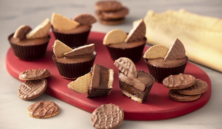 Foto de uma bancada branca. Sobre ela há uma tábua vermelha com seis unidades da receita pronta, além de alguns Biscoitos Calipso Cobertos com Chocolate.