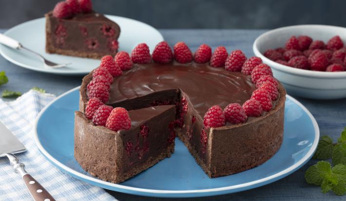 Fotografia em tons de vermelho em uma bancada de madeira azul, um pano azul quadriculado com uma espátula para bolo em cima dele. Ao centro, um prato azul redondo raso com a torta cremosa de chocolate com framboesas. Ao fundo, um prato com uma fatia.