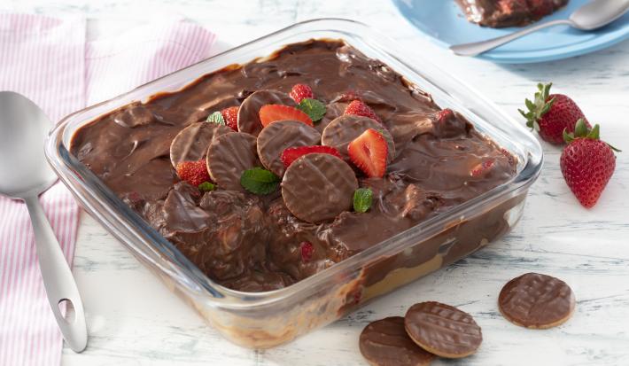 Fotografia em tons de vermelho em uma bancada de madeira clara, um pano branco com listras vermelhas, um refratário quadrado de vidro com a lasanha doce de chocolate com morangos e biscoito calipso dentro dele. Ao fundo, um prato azul com fatia do doce.