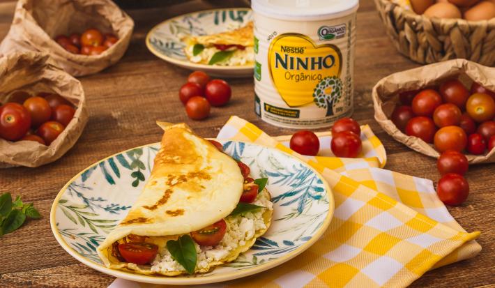 Fotografia em tons de amarelo e vermelho de uma bancada de madeira com um pano xadrez amarelo e branco, uma prato com uma crepioca recheada de queijo e tomate. Ao fundo tomates-cereja, uma lata de NINHO e ovos.