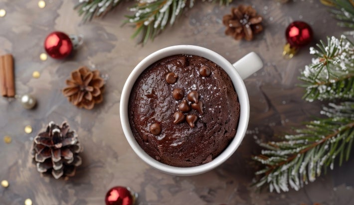 Caneca branca cheia de bolo de chocolate, decorada com gotas de chocolate e açúcar de confeiteiro. A caneca está cercada por enfeites natalinos, como ramos de pinheiro, pinhas e ornamentos vermelhos e prateados.