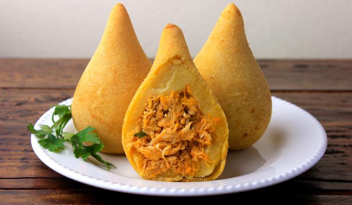 Fotografia em tons de branco com um prato branco ao centro. Em cima do prato existe duas coxinhas com massa de batata doce recheadas com frango