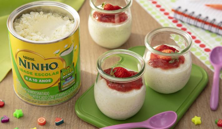 Fotografia de três recipientes de vidro pequenos com pudim de tapioca de ninho um creme de morango por cima. Dois desses recipientes estão sobre um apoio verde retangular, ao lado de uma colher roxa pequena e de uma lata de ninho escolar.