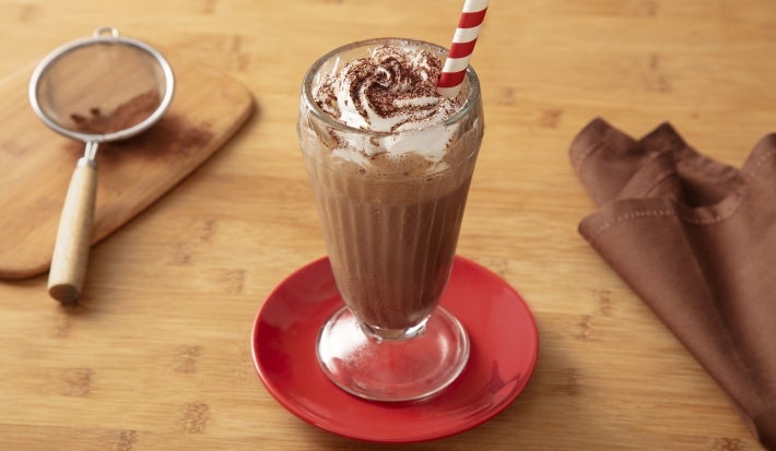 Fotografia em tons claros de um copo grande de vidro, de milkshake, com a bebida de nescau, finalizada com chantilly e nescau. O copo está sobre um mini prato vermelho, apoiado em uma mesa de madeira clara.