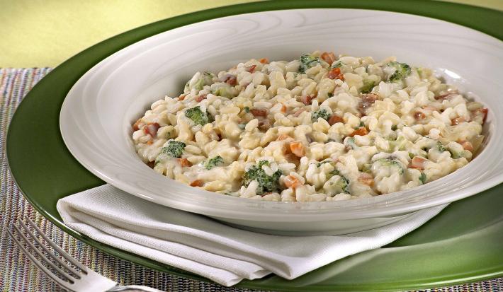 Fotografia em tons de verde em uma mesa de madeira com um jogo americano trançado colorido com um sousplat verde e um prato branco fundo com o risoto de legumes dentro dele.