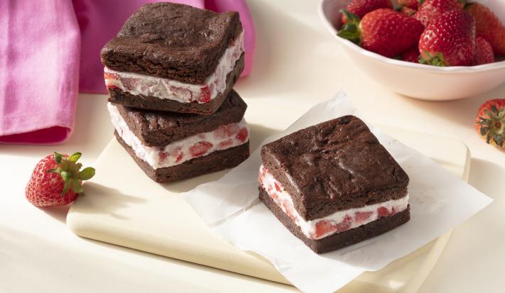 Imagem aproximada da receita de Sanduíche de Brownie com Sorvete, em quadrados marrons, recheados com um creme claro com pedaços de morango, todos sobre uma tábua clara em uma bancada decorada com mais morangos e um tecido em tom rosa